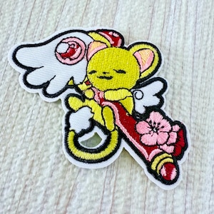 May include: An embroidered patch featuring a yellow creature with large ears and white wings, holding a red object adorned with a pink cherry blossom flower. The design includes a white border and black outlines, showcasing a stylized character.
