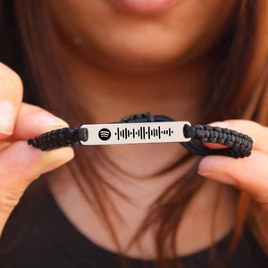 Puede incluir: Una pulsera trenzada negra con una placa rectangular plateada con un código de Spotify. La pulsera está sujeta por una persona. El logotipo de Spotify es visible en el lado izquierdo de la placa.