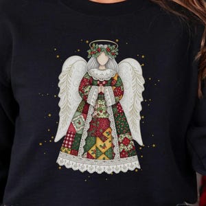 May include: Black sweatshirt featuring a detailed angel design with white wings, a floral halo, and a patchwork dress. The angel is centered on the chest, with gold star accents. The sweatshirt is paired with black and white checkered pajama pants.