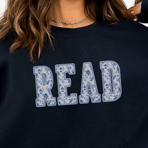May include: A navy blue sweatshirt featuring the word "READ" in large, light blue letters. The letters have a floral pattern. The sweatshirt is a casual style.