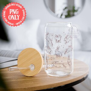 Peut inclure: Un verre à boire clair en forme de canette avec un couvercle et une paille en bambou. Le verre est décoré d'un motif de lapins esquissés. Le couvercle est brun clair. L'image comprend également un cercle rouge avec le texte "PNG ONLY".