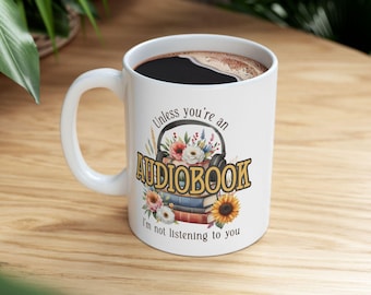 May include: White ceramic coffee mug with a black interior filled with coffee. The mug features the text "Unless you're an AUDIOBOOK I'm not listening to you" with a floral and headphone design. The mug is on a wooden surface.