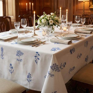 以下が含まれることがあります： 特別な日のためにセットされたダイニングテーブル。白いテーブルクロスには青い花の刺繍が施されています。テーブルには、お皿、カトラリー、水グラス、そして火の灯ったキャンドルがセットされています。白い花のアレンジメントが中央にあります。