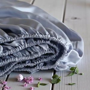May include: A stack of folded, light blue fitted sheets with elastic edges. The sheets are made of a silky material and are resting on a light gray wooden surface. Small pink flowers and green leaves are scattered around the sheets.