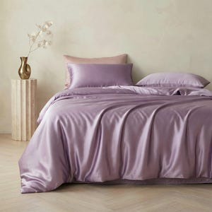 May include: A bed with a lavender silk duvet cover and pillowcases. The bedding has a lustrous sheen and is arranged on the bed. A light pink pillow and a gold vase with a decorative plant are also visible.