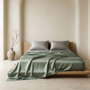 May include: A bed with sage green silk sheets and two gray pillowcases. The bed is on a light wood frame. A decorative vase with a branch is on the left side of the bed.