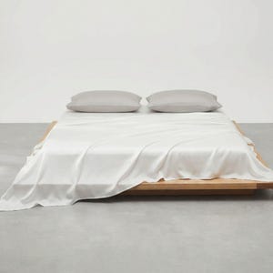 May include: A minimalist bedroom scene featuring a low wooden bed frame with a white sheet and two gray pillows. The bed is centered in the frame, with the sheet draped over the sides. The background is a plain white wall.