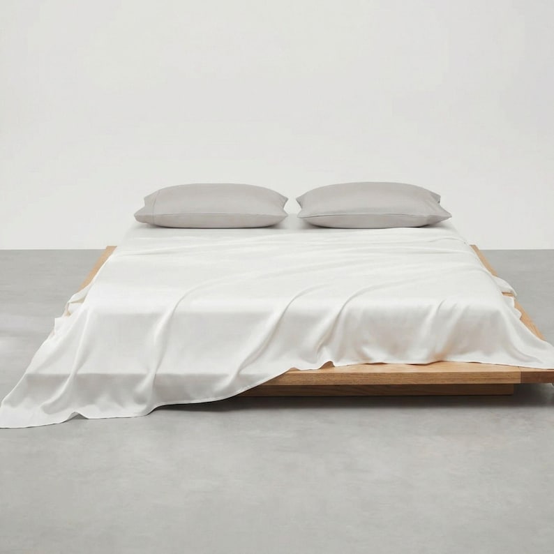 May include: A minimalist bedroom scene featuring a low wooden bed frame with a white sheet and two gray pillows. The bed is centered in the frame, with the sheet draped over the sides. The background is a plain white wall.