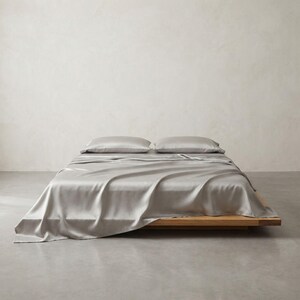 May include: A bed with a light gray silk sheet set and two matching pillows. The bed frame is made of light-colored wood. The background is a plain, off-white wall and a gray floor.