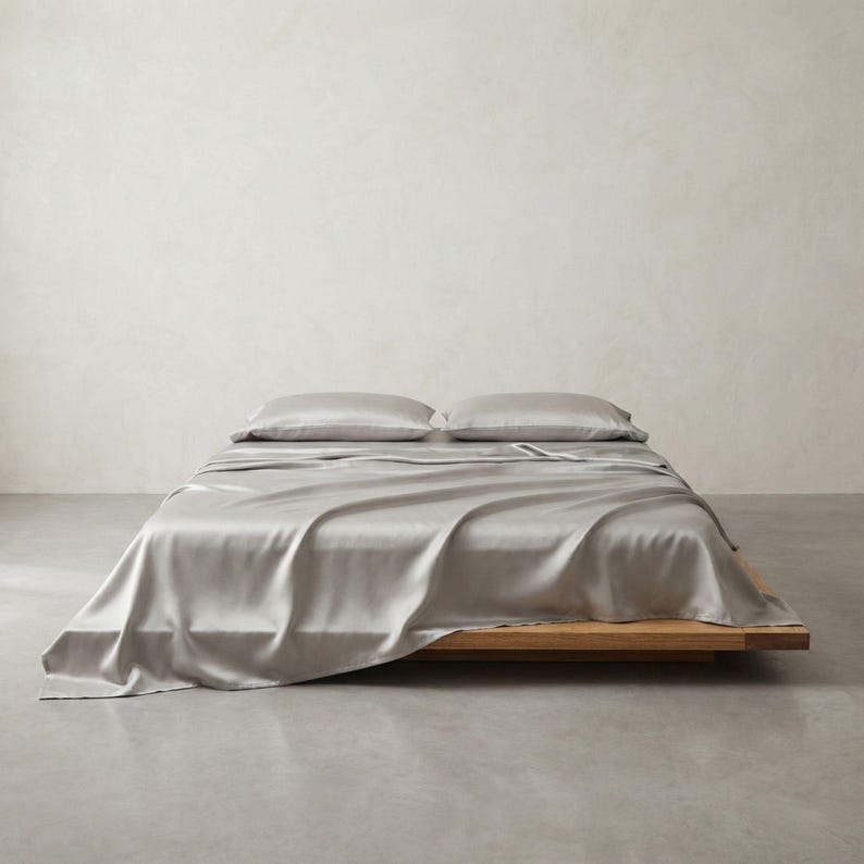 May include: A bed with a light gray silk sheet set and two matching pillows. The bed frame is made of light-colored wood. The background is a plain, off-white wall and a gray floor.
