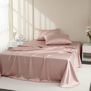 Op de afbeelding: Een bed met een blush roze satijnen lakenset. De set bevat een hoeslaken, een plat laken en twee kussenslopen. Het bed staat in een lichte kamer met een wit tapijt en een wit nachtkastje.