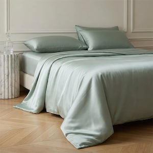May include: A bed with light green satin bedding, including a fitted sheet, duvet cover, and two pillowcases. The bed is neatly made, with the duvet cover draped over the side. A marble side table and a glass decanter are visible.