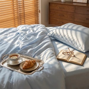 May include: A light blue bed with a matching duvet and pillow. A silver tray holds a cup of tea with a lemon slice and a croissant. An open book and eyeglasses rest on the bed, near a window with wooden blinds.