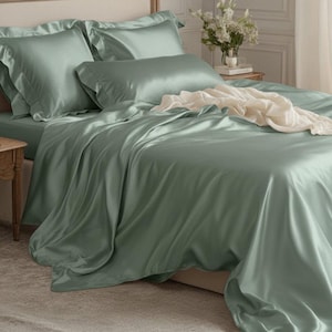 Peut inclure: Ensemble de lit en satin vert sauge, comprenant un drap-housse, des taies d'oreiller et une housse de couette. La literie a un éclat brillant et est disposée sur un lit avec une tête de lit crème. Un plaid crème est drapé sur le lit.