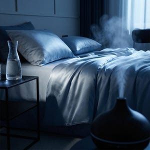 May include: A bedroom scene with a bed covered in light blue satin bedding. A black essential oil diffuser emits steam. A glass carafe of water sits on a black side table next to the bed. The room is dimly lit, creating a relaxing atmosphere.
