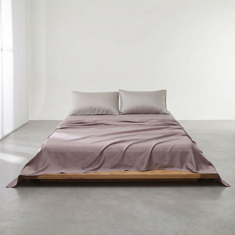 May include: A bed with a wooden frame, covered with a mauve-colored sheet set and two matching pillowcases. The bed is in a minimalist room with a white wall and a gray floor.