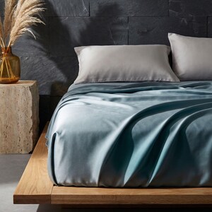 May include: A bed with a light blue sheet set and two gray pillows. The bed frame is made of light-colored wood. A vase with dried plants sits on a stone side table. The background is a dark gray stone wall.