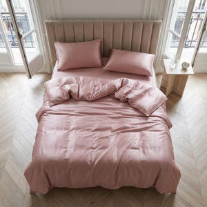 May include: A bed with a pink satin bedding set, including a duvet cover, fitted sheet, and two pillowcases. The bed has a padded headboard and is placed in a room with large windows and a wooden floor.