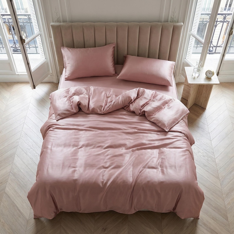 May include: A bed with a pink satin bedding set, including a duvet cover, fitted sheet, and two pillowcases. The bed has a padded headboard and is placed in a room with large windows and a wooden floor.