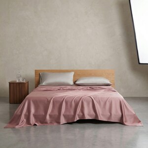 May include: A bed with a wooden headboard is covered with a dusty rose-colored sheet set. Two gray pillows rest on the bed. A small wooden nightstand with a glass carafe and two glasses sits to the left of the bed.