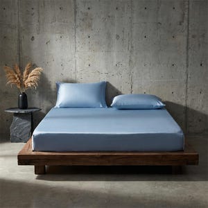 May include: A light blue bed set, including a fitted sheet and two pillowcases, on a wooden bed frame. The bedding has a smooth, satin-like finish. A black vase with dried plants sits on a small side table.