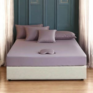 May include: A bed with a lavender-colored fitted sheet, matching pillowcases, and a sleep mask. The bed is made up with a white base and is set against a teal wall with white curtains on either side.