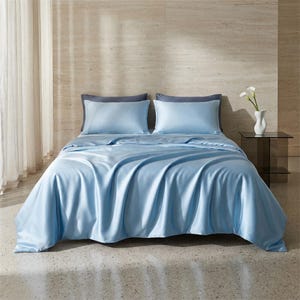 May include: A bed is made with light blue satin bedding, including a duvet cover and two pillowcases. The pillowcases are a darker shade of blue. The bed is in a room with a neutral-colored wall and a small glass side table.