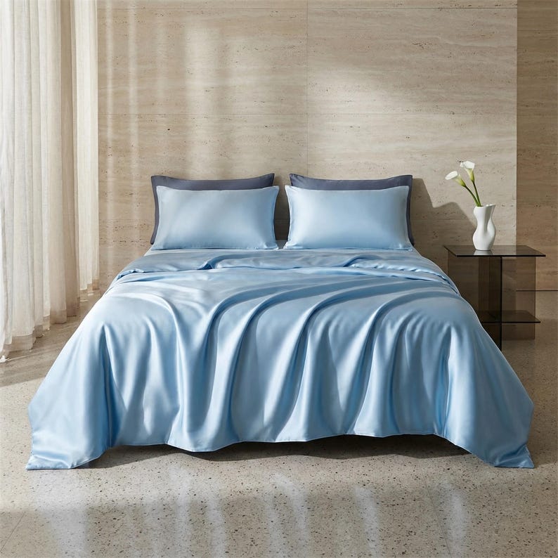 May include: A bed is made with light blue satin bedding, including a duvet cover and two pillowcases. The pillowcases are a darker shade of blue. The bed is in a room with a neutral-colored wall and a small glass side table.