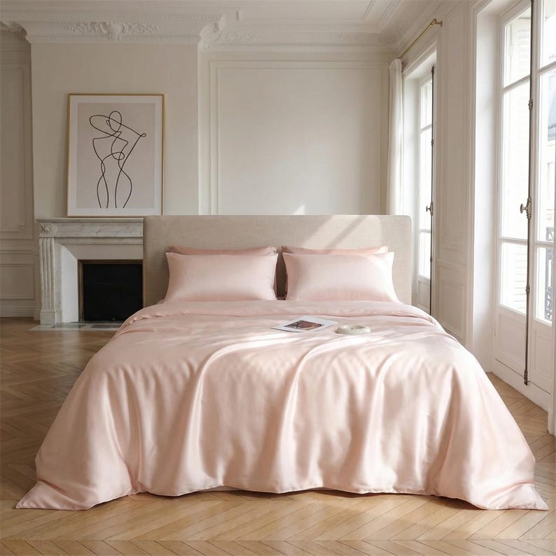 May include: A bed with light pink satin bedding, including a duvet cover and two pillowcases. The bed is made up and ready for use. The room has a neutral color palette with a framed art print and a fireplace.