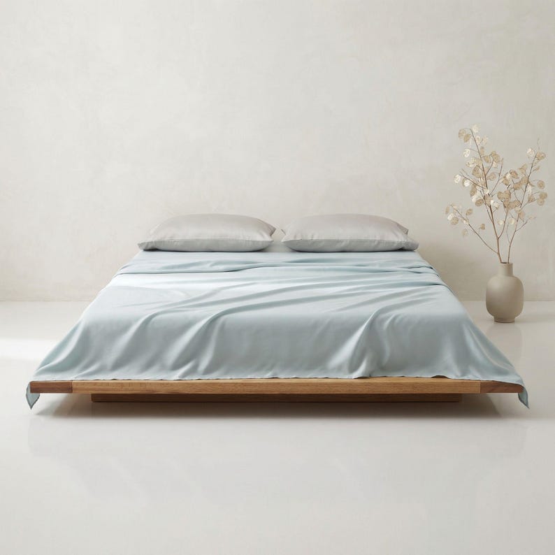 May include: A bed with light blue sheets and two gray pillows. The bed frame is made of wood. A vase with dried flowers sits to the right of the bed. The background is a light, neutral color.
