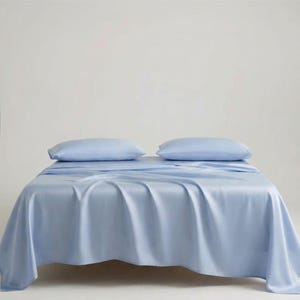 May include: A bed with light blue bedding, including a fitted sheet, flat sheet, and two pillowcases. The sheets are draped over the bed, creating soft folds. The background is a plain white.
