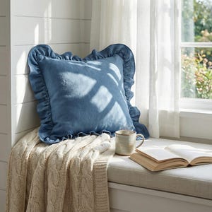 May include: A blue cushion with a frilled border sits on a cream cable-knit throw. A ceramic mug and an open book are placed on a window seat, creating a cosy reading area.