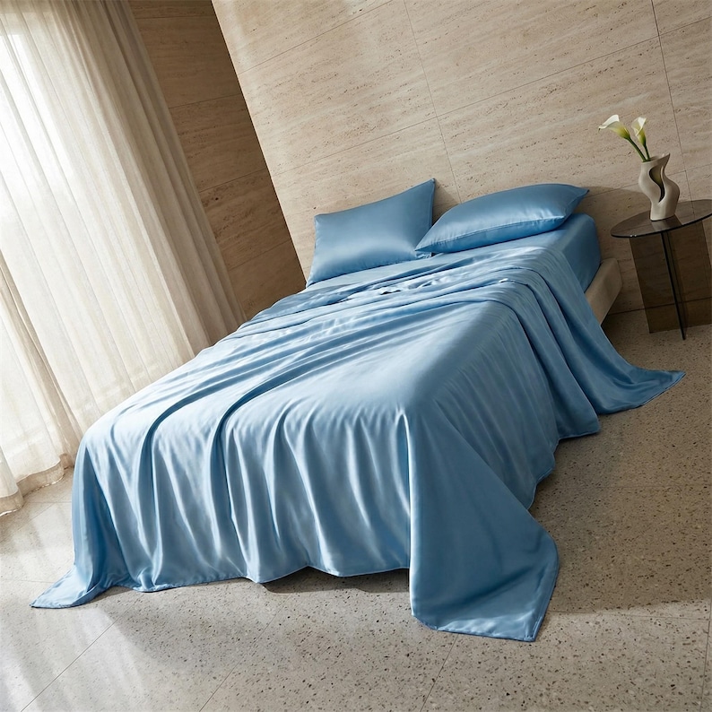 May include: A bed is covered with light blue satin sheets and pillowcases. The sheets drape over the sides of the bed. The room has a neutral color palette with a window and a small table with a vase of flowers.