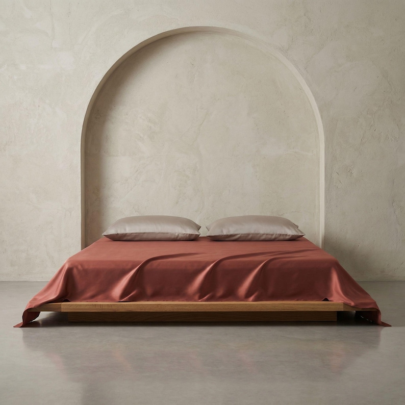 May include: A modern bedroom setup featuring a low wooden bed frame with a terracotta-colored silk sheet. Two light gray pillows rest on the bed. The background includes a neutral-toned wall with an arched alcove.