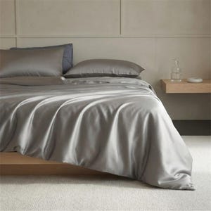 May include: A bed with gray satin bedding, including a duvet cover and two pillows. The bed is made up with a gray sheet set. The bed is on a wooden frame. A small wooden side table is next to the bed.