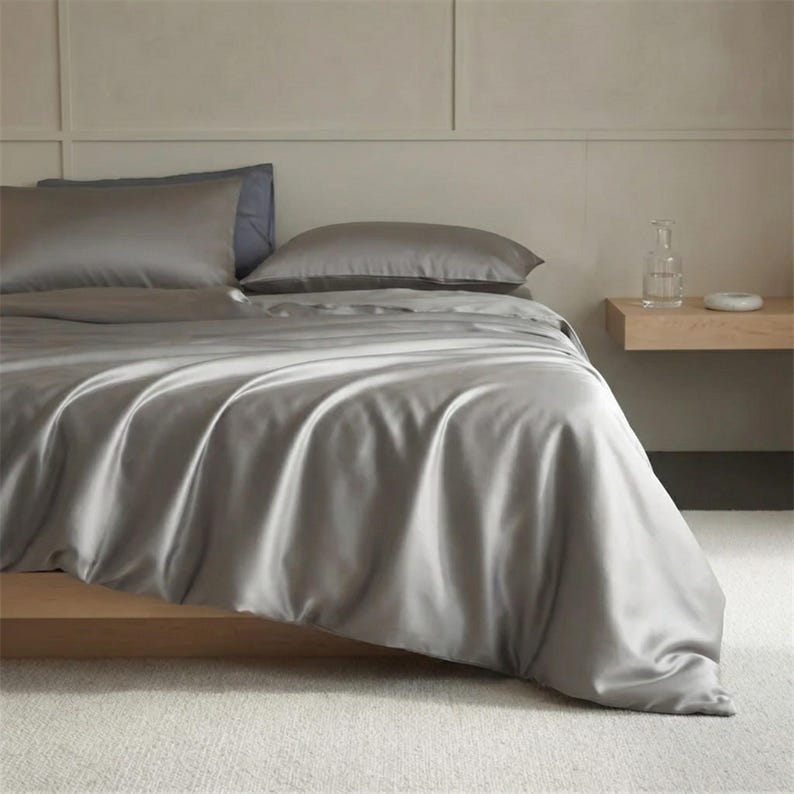 May include: A bed with gray satin bedding, including a duvet cover and two pillows. The bed is made up with a gray sheet set. The bed is on a wooden frame. A small wooden side table is next to the bed.