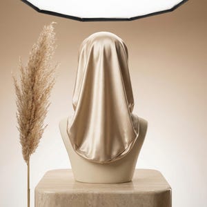 Puede incluir: Un hiyab de satén dorado drapeado sobre una cabeza de maniquí. El hiyab es liso y brillante, reflejando la luz. A la izquierda del maniquí hay una hierba ornamental alta y seca. El fondo es de color beige suave.