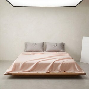 May include: A bed with a light pink sheet set and two gray pillowcases. The bed frame is made of wood. The room has a neutral color palette with a white wall and a gray floor.