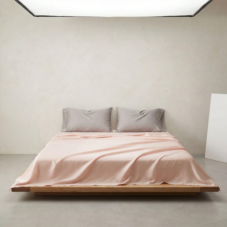 May include: A bed with a light pink sheet set and two gray pillowcases. The bed frame is made of wood. The room has a neutral color palette with a white wall and a gray floor.