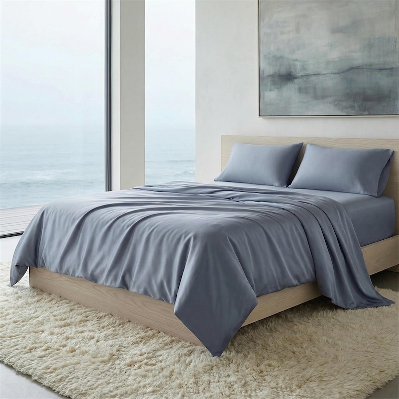 May include: A bed with a light wood frame is covered with a set of blue-grey sheets and pillowcases. The bed sits on a cream-colored, shaggy rug. A large window and a painting are visible in the background.