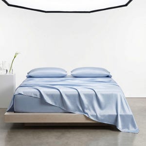May include: A light blue bed set, including a fitted sheet, flat sheet, and two pillowcases, is displayed on a wooden bed frame. The sheets have a silky, smooth texture. The bed is in a bright, minimalist room with a white wall and a concrete floor.