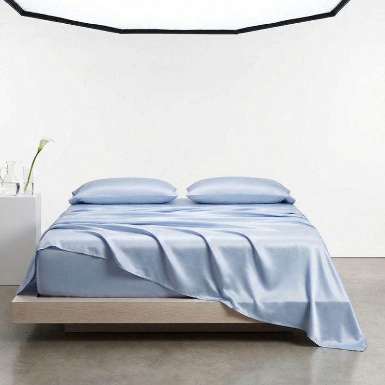 May include: A light blue bed set, including a fitted sheet, flat sheet, and two pillowcases, is displayed on a wooden bed frame. The sheets have a silky, smooth texture. The bed is in a bright, minimalist room with a white wall and a concrete floor.