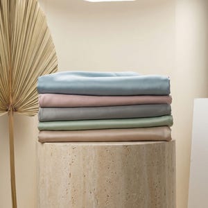 May include: A stack of six folded fabric sheets in pastel colors: light blue, pink, gray, green, and tan. The sheets are neatly arranged on a cylindrical stone pedestal. A large, dried palm leaf is visible in the background.