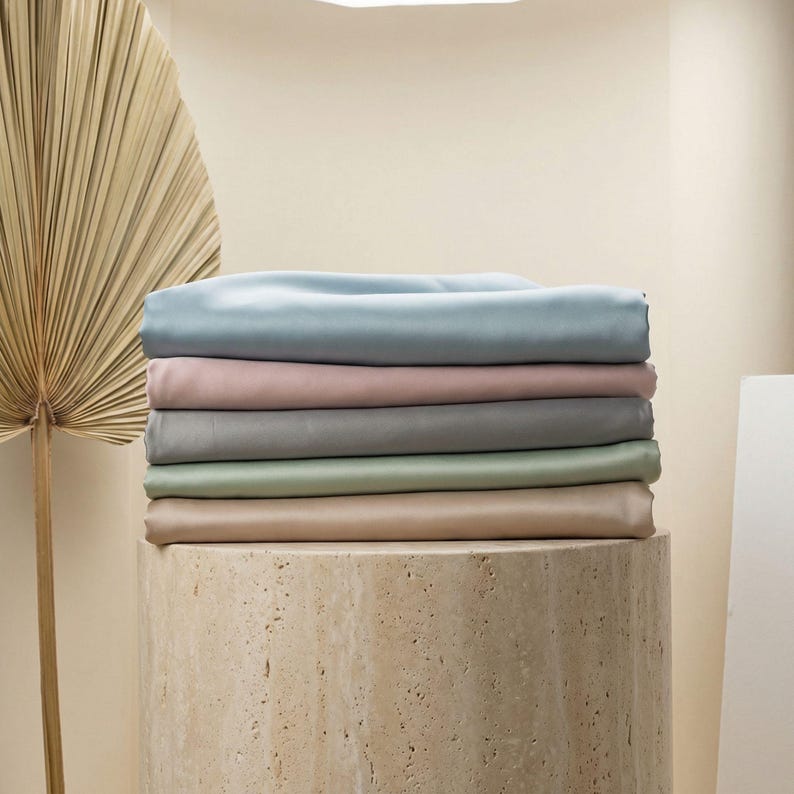 May include: A stack of six folded fabric sheets in pastel colors: light blue, pink, gray, green, and tan. The sheets are neatly arranged on a cylindrical stone pedestal. A large, dried palm leaf is visible in the background.