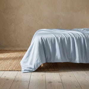 May include: A bed covered with a light blue sheet. The sheet drapes over the side of the bed, revealing a glimpse of a woven rug on the floor. The floor is made of light-colored wood.
