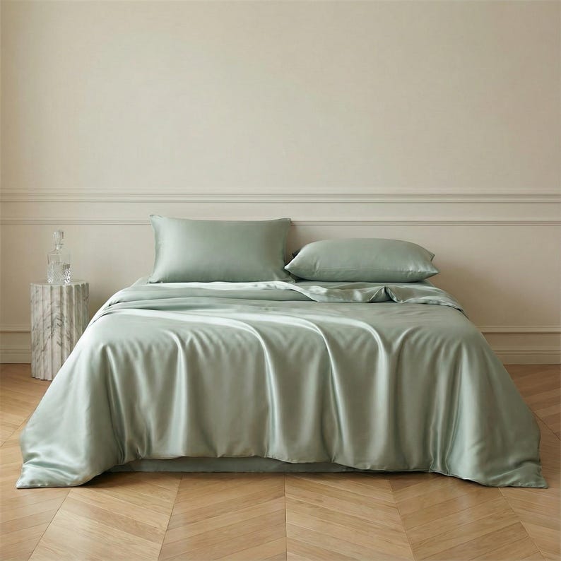 May include: A bed with sage green silk bedding. The set includes a duvet cover and two pillowcases. The bed is made up with a fitted sheet and a flat sheet. The room has a neutral color palette with a marble side table.