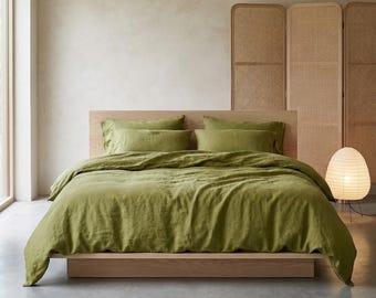Op de afbeelding: Een bed met olijfgroene beddengoed, inclusief een dekbedovertrek en kussenslopen. Het bedframe is van lichtgekleurd hout. Een geweven kamerscherm en een papieren lantaarn staan op de achtergrond, wat een minimalistische slaapkamer creëert.