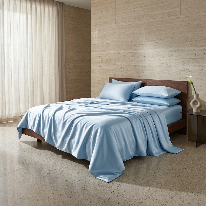 May include: A bed with light blue bedding, including a fitted sheet, flat sheet, and pillowcases. The bed has a dark wooden frame and is set against a neutral-toned wall. A small vase with a white flower sits on a side table.