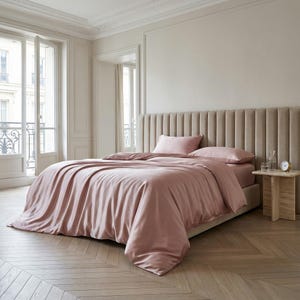 May include: A bed with a blush pink satin duvet cover and matching pillowcases. The headboard is a light beige, vertically ribbed design. The room has light wood flooring and a small side table with a glass of water and a clock.