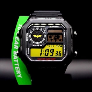 May include: Black digital watch with a yellow display showing the time and date. The watch features a Batman logo and the text "10 YEAR BATTERY". A green tag with "10 YEAR BATTERY" is attached.